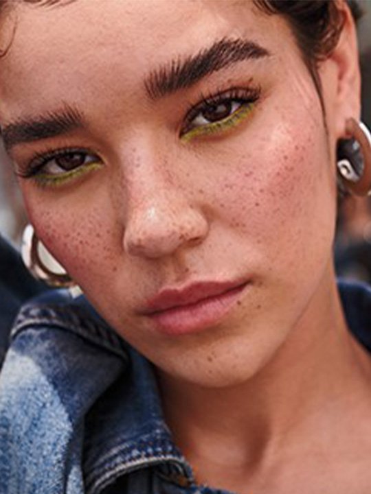 Nahaufnahme einer Frau mit auffälligem grünem Eyeliner unter dem Auge, frechem Ausdruck und Sommersprossen – urbaner, gewagter Look.