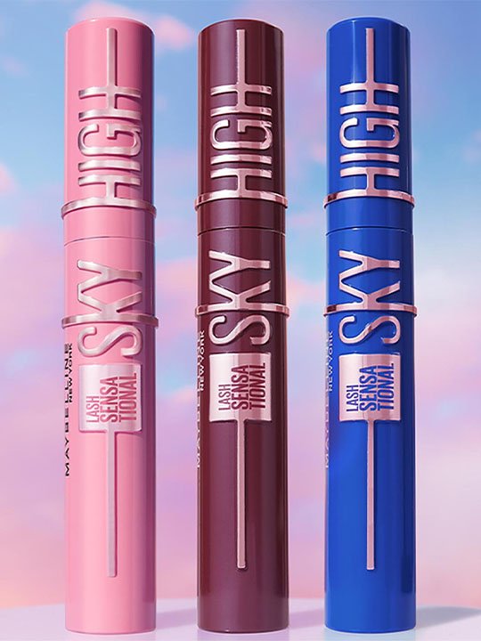 Drei Maybelline Lash Sensational Sky High Mascaras in Rosé, Bordeaux und Blau vor pastelligem Himmel – trendige Farbvarianten für individuelle Looks.