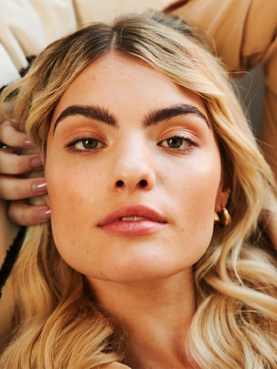 Blondes Model mit natürlichen Brauen und dezentem Make-up in entspannter Pose.