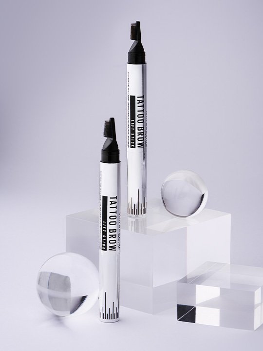 Zwei Maybelline Tattoo Brow Lift Stifte auf minimalistischem, modernem Untergrund mit transparenten geometrischen Objekten.