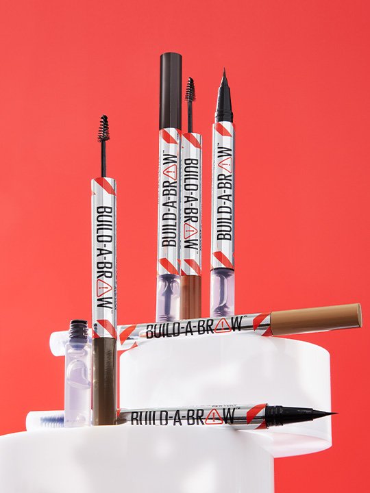 Build-A-Brow Augenbrauenprodukte in verschiedenen Farben und Applikator-Varianten vor kräftigem roten Hintergrund inszeniert.