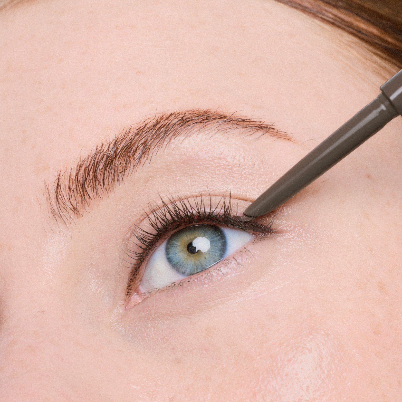 Nahaufnahme eines Auges, an dessen oberen Wimpernkranz ein brauner Eyeliner gezogen wird