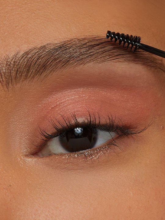 Nahaufnahme einer geschminkten Augenpartie mit einer Brauenbürste, die die Augenbraue formt – natürlicher Look mit dezentem Lidschatten.