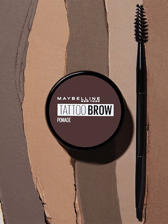 Tattoo Brow Pomade von Maybelline New York neben einer Brauenbürste, auf Texturflächen in verschiedenen Brauntönen präsentiert.