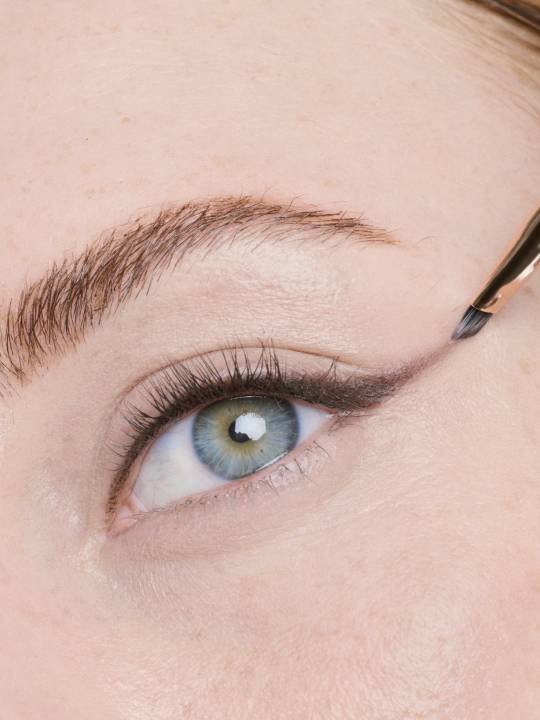 Nahaufnahme einer jungen Frau mit blauen Augen, die sich mit einem kleinen Pinsel Kajal aufträgt