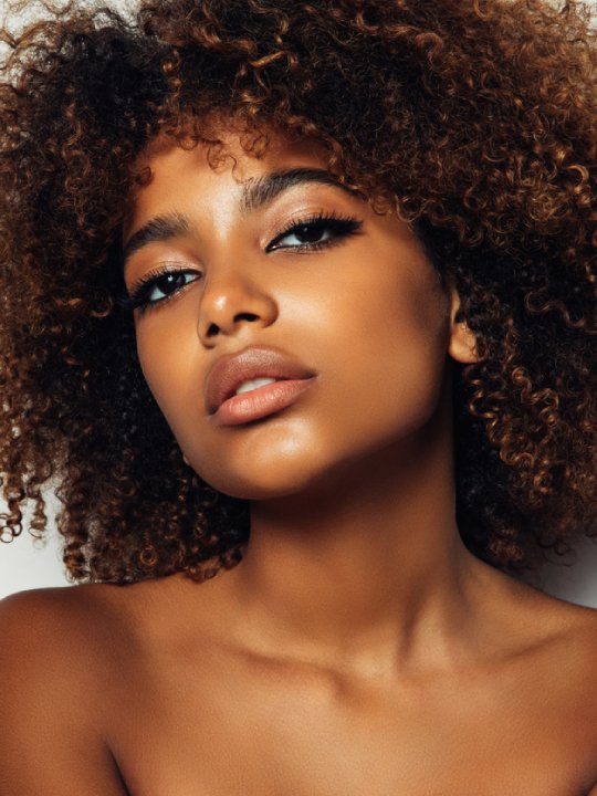 Porträt einer Frau mit lockigem braunem Haar, warmem Hautton, dezentem Make-up und intensivem Blick vor neutralem Hintergrund.