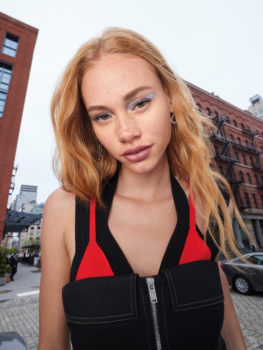 Porträt einer jungen Frau mit roten Haaren, Sommersprossen und auffälligem Make-up in urbaner Umgebung, umgeben von Backsteingebäuden.