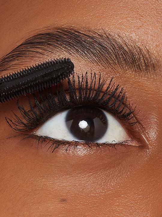 Nahaufnahme eines geschminkten Auges mit dunkler Iris, während eine Mascara-Bürste die oberen Wimpern tuscht.
