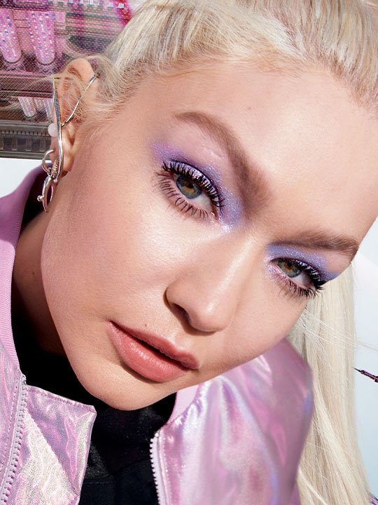 Model mit platinblondem Haar, intensivem lila Lidschatten und voluminösen Wimpern blickt in die Kamera.