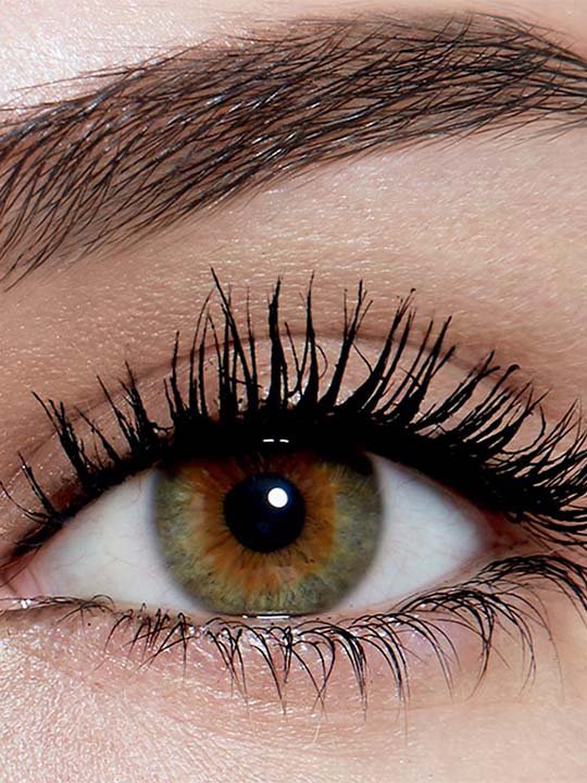 Makroaufnahme eines Auges mit grün-brauner Iris und stark getuschten, voluminösen Wimpern.
