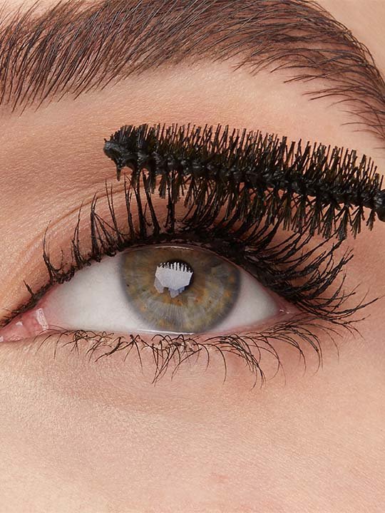 Makroaufnahme eines Auges mit hellgrauer Iris – die oberen Wimpern werden gerade mit einer schwarzen Mascara-Bürste getuscht.