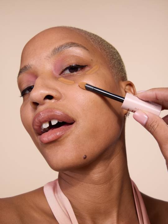 Nahaufnahme einer jungen Frau mit kurz-rasierten Haaren, die Concealer unter ihrem Auge aufträgt