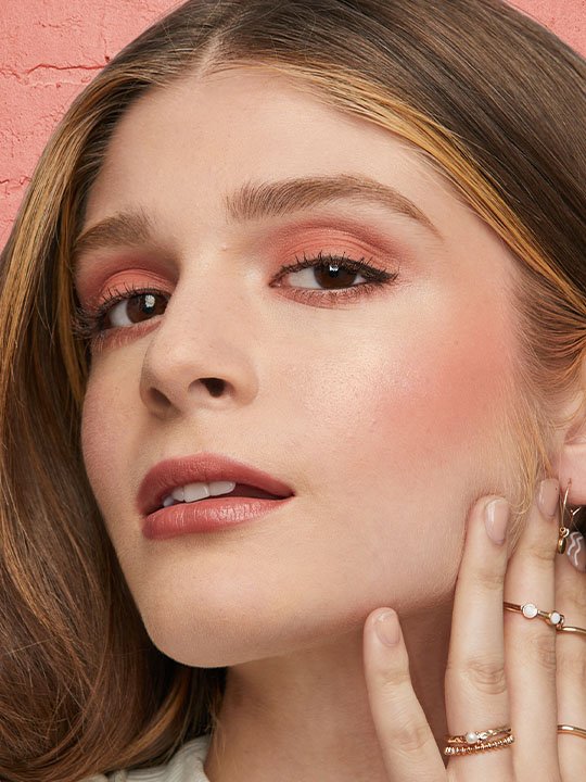 Porträt einer Frau mit warmem Pfirsichton auf Augen und Wangen sowie dezentem Lippenstift vor strukturierter rosa Wand.