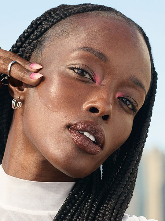 Stilvolles Porträt einer Frau mit Flechtfrisur, dunklem Lippenstift und pinkem Lidschatten, die sich das Gesicht leicht berührt.