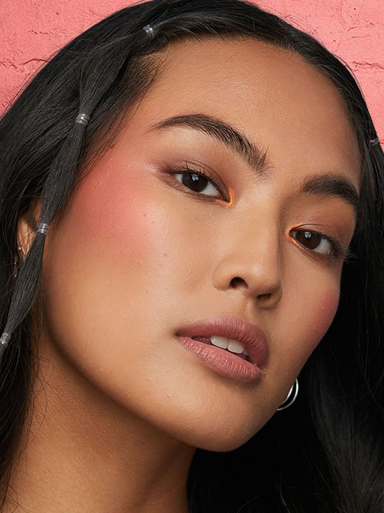 Frau mit frischem Make-up-Look, auffälligem orangefarbenem Lidstrich im Augeninnenwinkel und betonten Wangen.