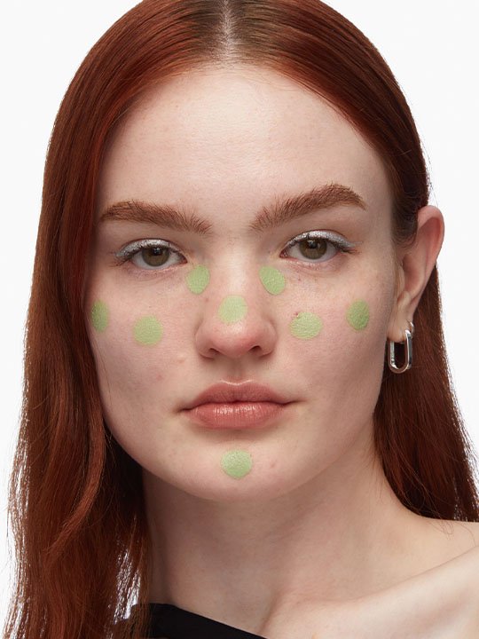 Beauty-Anwendung: Rotehaarige Frau mit grünen Farbkorrigierpunkten auf Stirn, Wangen, Nase und Kinn vor weißem Hintergrund.