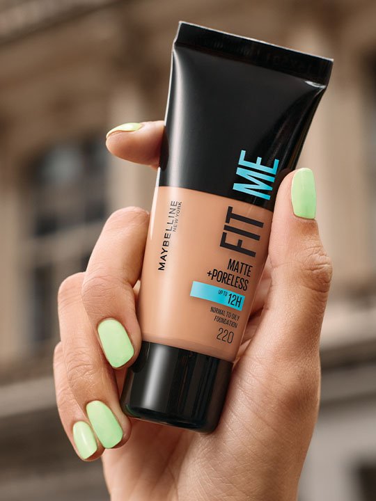 Hand hält Maybelline Fit Me Foundation vor unscharfem Stadt-Hintergrund.