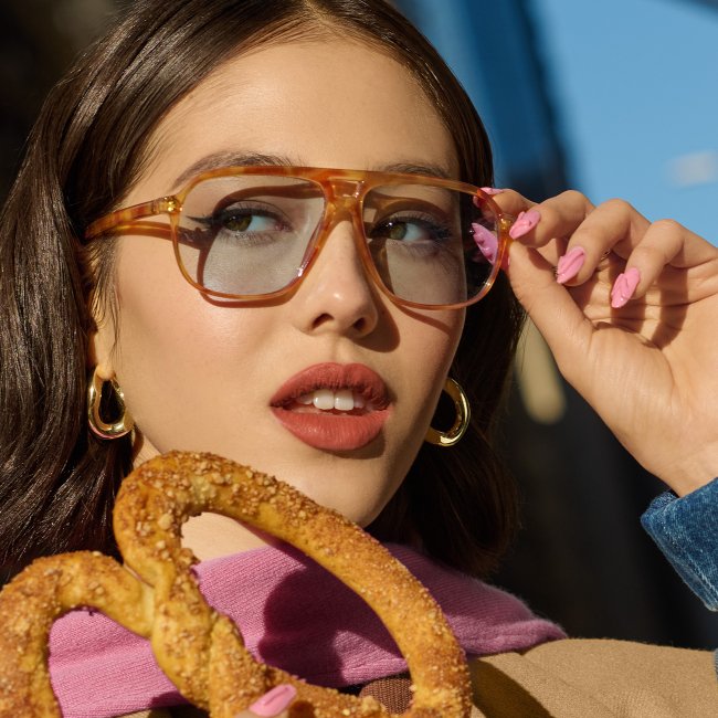 Junge Frau mit langen, braunen Haaren und einer modischen, orangenen Sonnenbrille hält eine Brezel