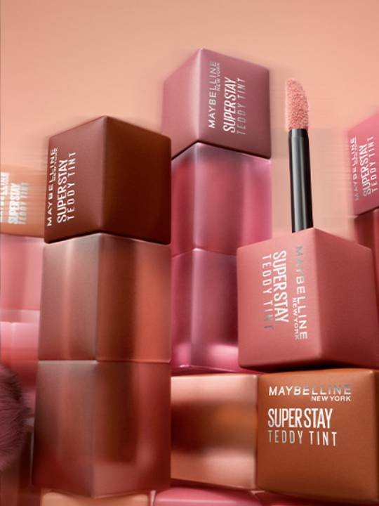 Mehrere Maybelline Teddy Tint Lippenstifte in verschiedenen Farben vor einem pfirsichfarbenen Hintergrund