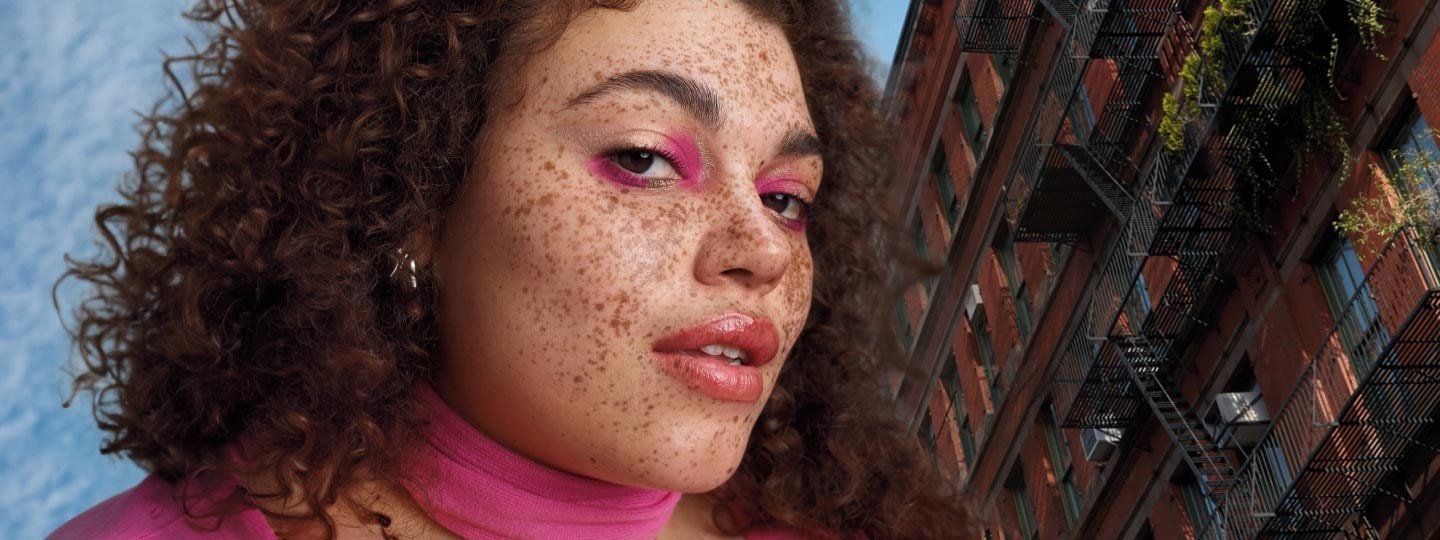 Eine Frau mit Sommersprossen und pinken Augen Make-Up steht vor einem Gebäude.