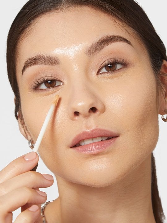 Frau trägt Concealer mit Applikator unter dem Auge auf, um Augenschatten optisch zu kaschieren.