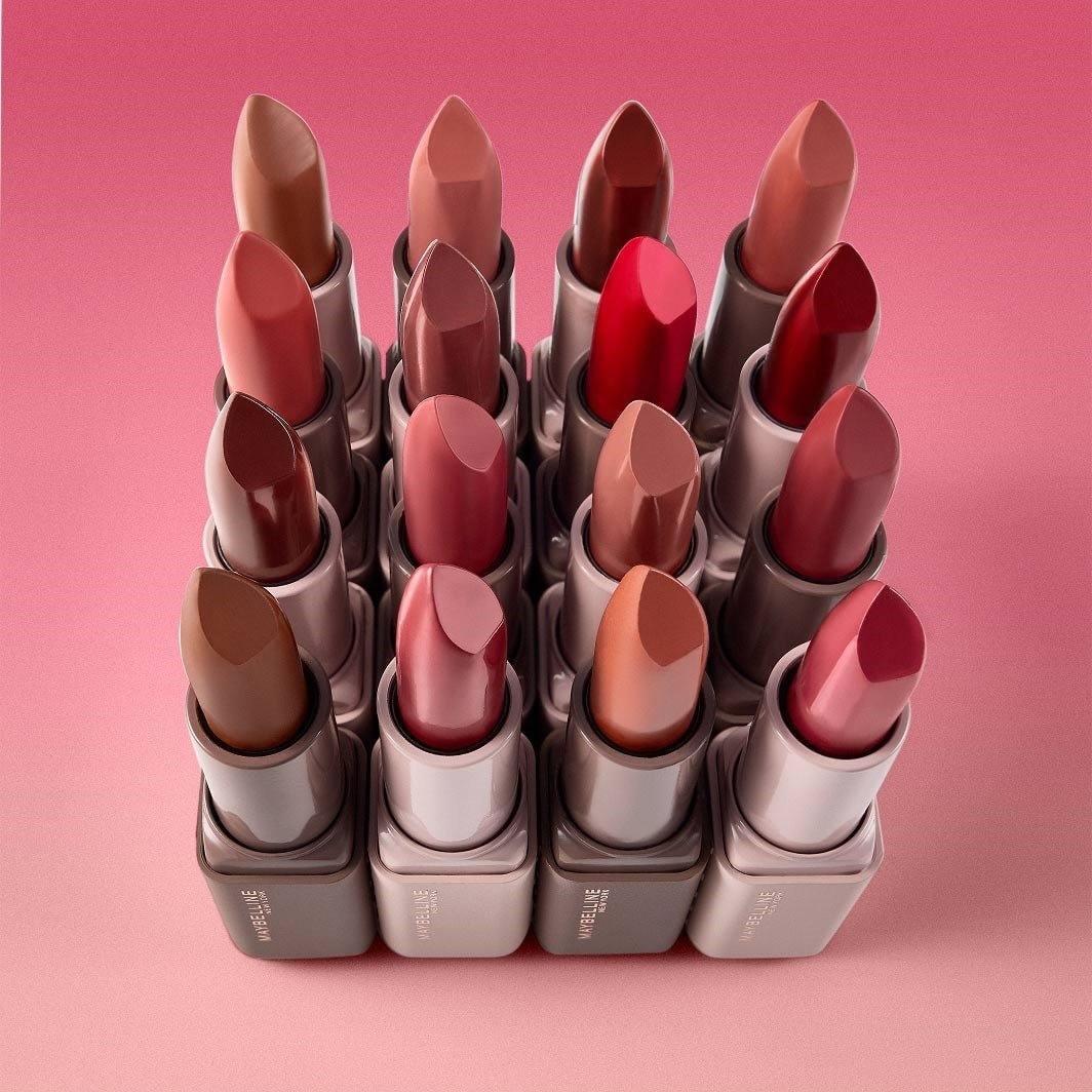 Nahaufnahme eines Rasters von Maybelline Lippenstiften in verschiedenen Nude-, Rosa-, Rot- und Brauntönen, die auf einem rosa Hintergrund angeordnet sind.
