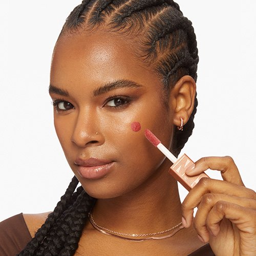 Frau hält MAYBELLINE New York Sunkisser Blush in der Hand und hat 1 Punkt aus Blush auf ihrer Wange. 
