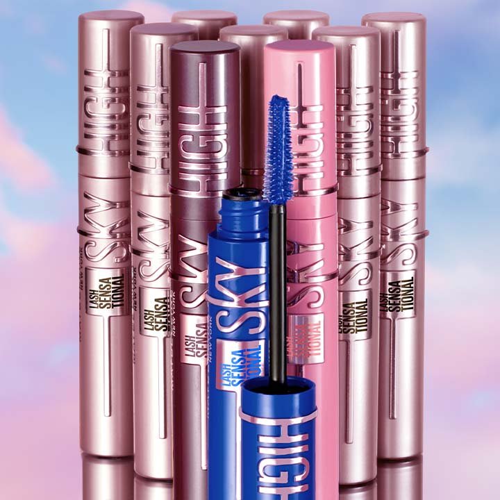 Mehrere Maybelline Lash Sensational Sky High Mascaras stehen nebeneinander, im Vordergrund eine geöffnete blaue Mascara mit sichtbarer Bürste – präsentiert vor einem pastellfarbenen Himmel.