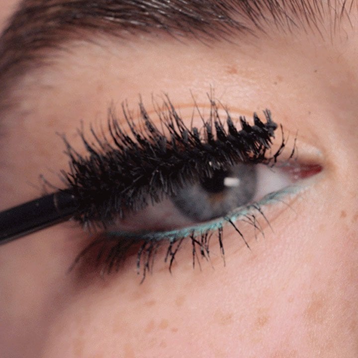 Nahaufnahme eines Auges mit blauen Augen, während schwarze Mascara auf die oberen Wimpern aufgetragen wird. Unter dem Auge ist türkisfarbener Eyeliner sichtbar.