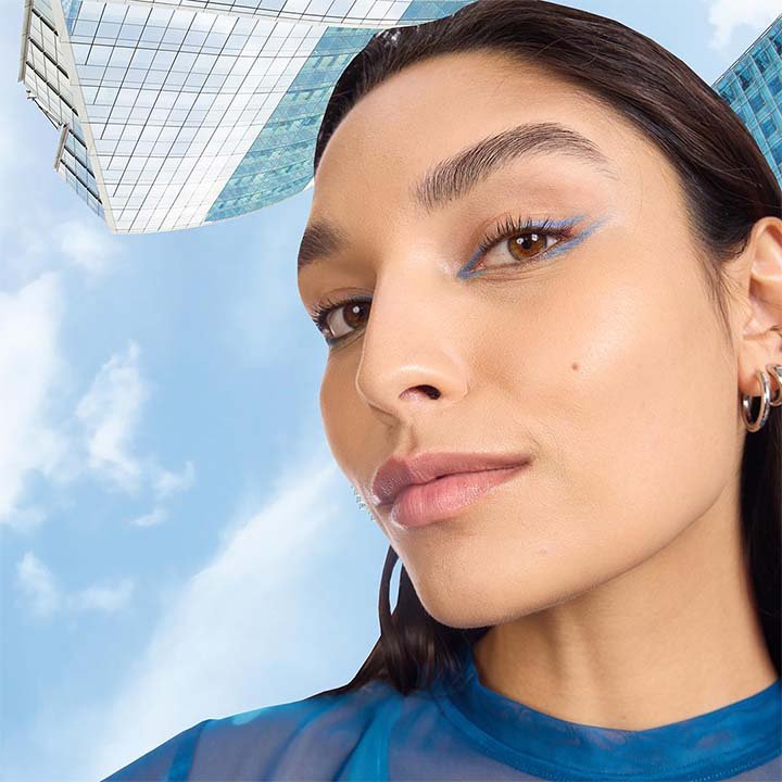 Nahaufnahme einer Frau mit markantem blauem Eyeliner und glattem Haar vor einem Himmel- und Gebäude-Hintergrund.