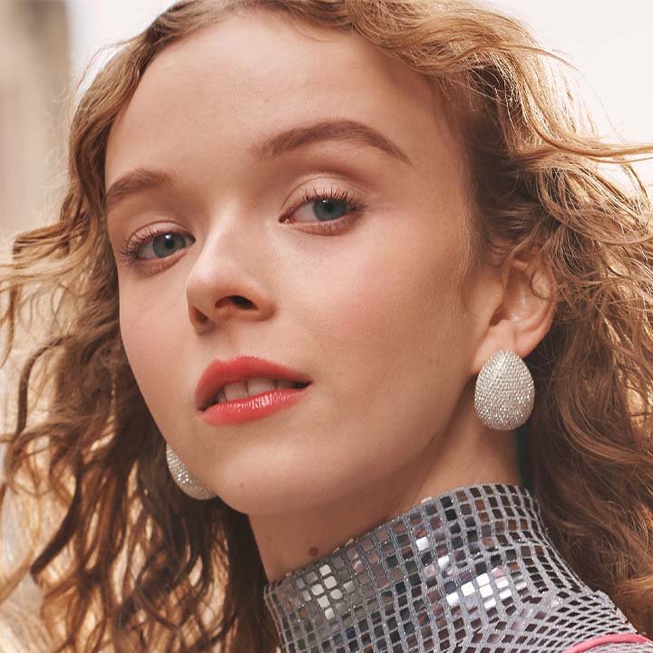 Model mit roten Lippen, glitzernden Ohrringen und silbernem Pailletten-Top blickt selbstbewusst in die Kamera.