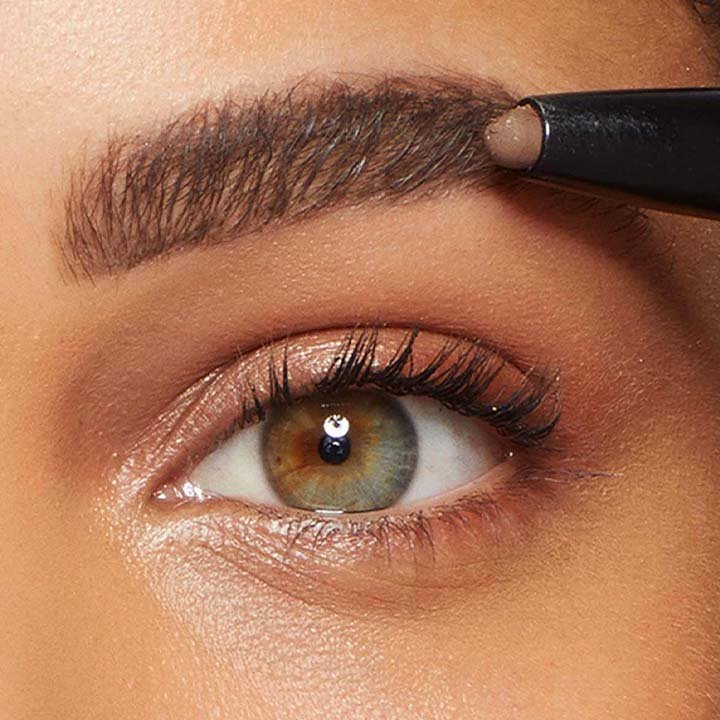 Nahaufnahme eines Auges mit grün-brauner Iris, während ein Brauenstift präzise die Augenbraue nachzeichnet – Fokus auf das Styling und Definieren der Augenbraue.