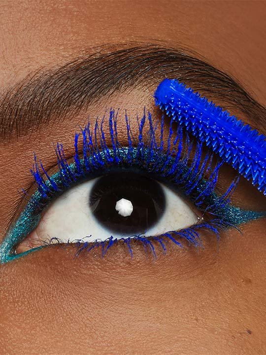 Makroaufnahme eines Auges mit türkisfarbenem Lidstrich und intensiv blauer Mascara, die mit einer passenden Bürste aufgetragen wird.