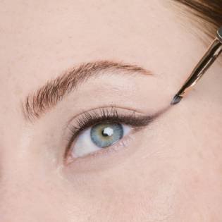 Nahaufnahme einer jungen Frau mit blauen Augen, die sich mit einem kleinen Pinsel Kajal aufträgt