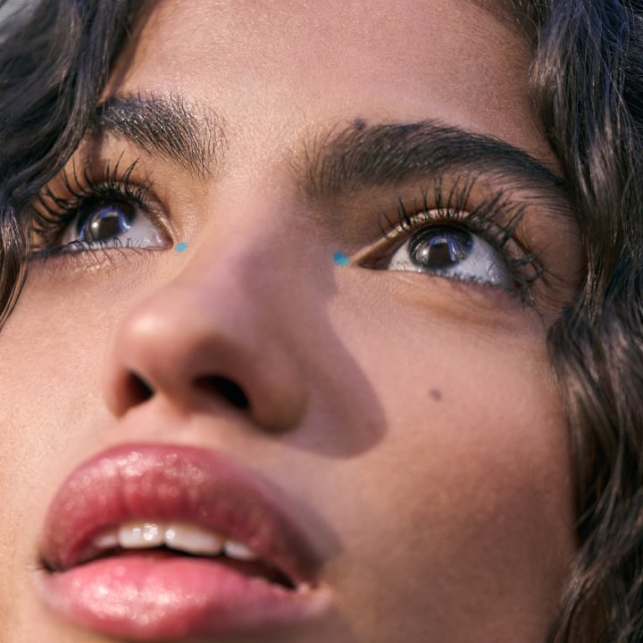 Nahaufnahme eines weiblichen Gesichts mit glänzenden Lippen, definierten Wimpern und kleinen blauen Akzenten im inneren Augenwinkel.